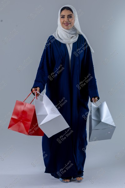 A portrait of a woman wearing a blue abaya and a white hijab, holding shopping bags in various colors - red, white, and silver. The woman stands against a light grey background and appears to be smiling.
