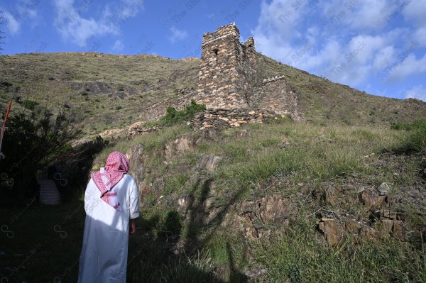 A shot of a Saudi man wearing the Saudi dress, meditating and standing on one of the green mountains.