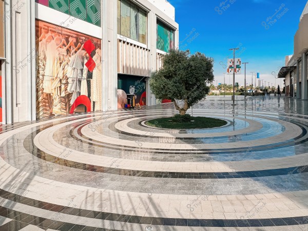 The image shows the facade of a modern shopping center, featuring a large fashion display with multi-colored dresses. The ground has a circular gradient tile design in black and white. In the center, there is a small green tree planted within a grassy circle. The background shows a clear sky with several street lamps lined along the path.
