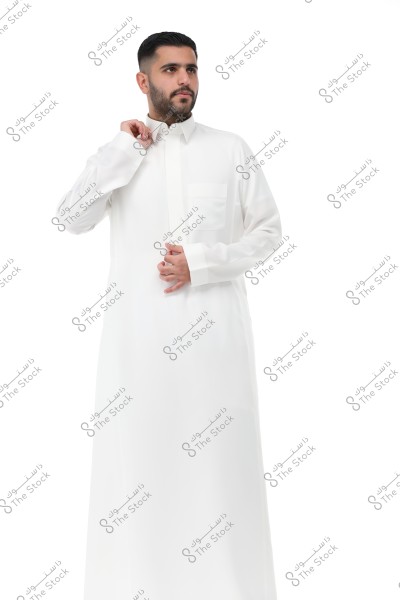 An image of a man wearing a white thobe, standing against a white background. The man is holding the collar of the thobe, appearing to adjust it. The thobe represents traditional Saudi Arabian attire.