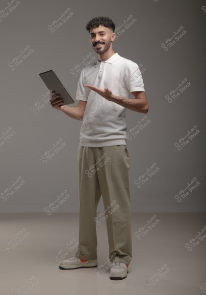 A portrait of a man standing and smiling while holding a tablet in his left hand. The man is wearing a white polo shirt, light brown trousers, and white sneakers. The background is a simple grey, suggesting the setting is a photo studio.