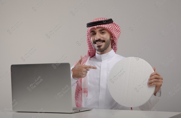 An image of a man sitting behind a gray laptop, wearing traditional Saudi attire with a red checkered headscarf (ghutra) and black agal. He is holding a round white board and pointing at it with his finger. The background is white, giving the image a simple look.