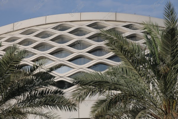 An image of a building with a contemporary architectural design featuring a facade adorned with wave-like repetitive patterns. In the foreground, green palm trees add a natural touch to the scene, with a clear sky in the background.