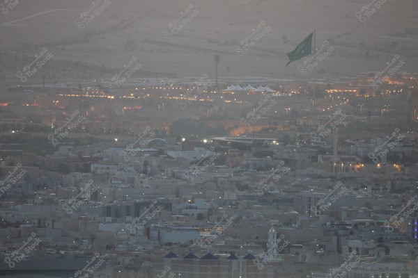 منظر جوي لمدينة سعودية عند الغروب, يظهر فيها مبانٍ سكنية ذات تصميم حديث, وأعمدة إنارة تزين الطرق. في الخلفية, يظهر علم كبير للمملكة العربية السعودية يرفرف, ومناطق مضيئة توحي بوجود منشآت وحدائق عامة.