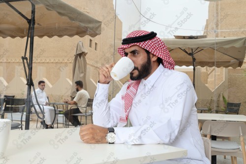 رجل سعودي يرتدي الزي السعودي التقليدي يضع ساعة على معصمه بينما يحتسي كوب من القهوة أثناء النهار