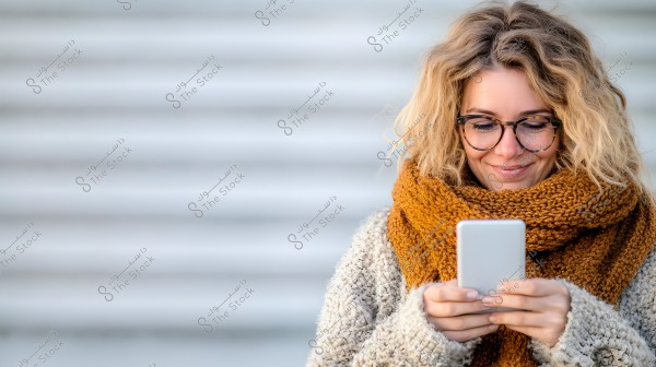 A woman smiling as she looks at her smartphone. She is wearing glasses and has wavy blonde hair. She is dressed in a large brown scarf and a gray knitted sweater. The background is blurry with horizontal lines.