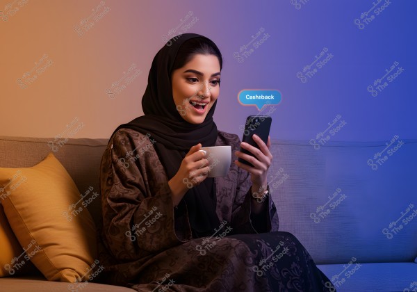 The image shows a woman wearing a black abaya and headscarf sitting on a grey sofa, holding a smartphone in her left hand and a coffee cup in her right hand. She appears happy as she looks at the phone, which displays a notification with the word \"Cashback\". The sofa is adorned with yellow cushions, and the warm lighting creates a cozy ambiance.