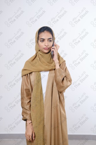 Portrait of a Saudi girl wearing an abaya on a white background holding a phone and making gestures with her hands while smiling, souvenir photos, documenting a happy moment.