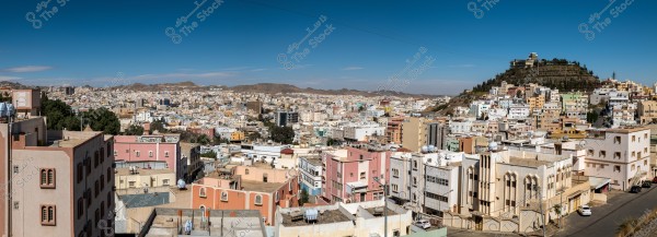 Panoramic view of a city with low-rise, colorful buildings stretching across a mountainous landscape. The buildings vary in shades of beige, pink, and other light colors. In the background, there is a hill topped with trees and a structure. The sky is blue and clear.
