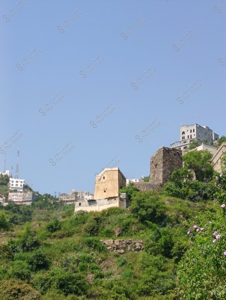 A landscape showing traditional buildings on green hills under a clear blue sky. The hills have lush vegetation and a stone tower next to another beige building. There are several modern buildings in the background on other hills, with radio antennas visible.