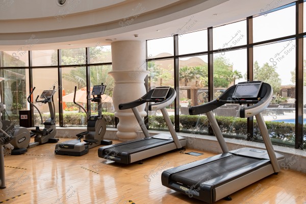 تُظهر الصورة صالة رياضية مع أجهزة مشي حديثة تطل على حديقة خارجية. تُحيط بالنوافذ الزجاجية الكبيرة التي تسمح بدخول الكثير من الضوء الطبيعي. الأرضيات مصنوعة من الخشب المصقول, وهناك نباتات وزهور خارج النوافذ تضفي جواً مريحاً.