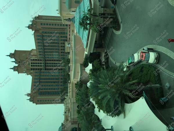 The image depicts Atlantis Hotel in Dubai, showcasing its complete architectural design, including pointed towers and tall windows. The foreground features the hotel\'s entrance with several cars parked alongside the curbs. The surrounding area includes palm trees and greenery, with some white canopies visible in the bottom part of the image. The sky is clear, providing a stunning view of the large structure.