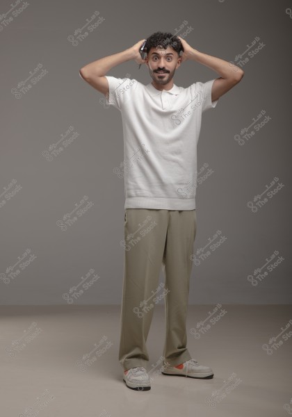 A portrait of a man standing with his hands on top of his head, smiling. He is wearing a white polo shirt, loose beige pants, and sneakers. The background is gray and neutral. The attire appears casual and comfortable.