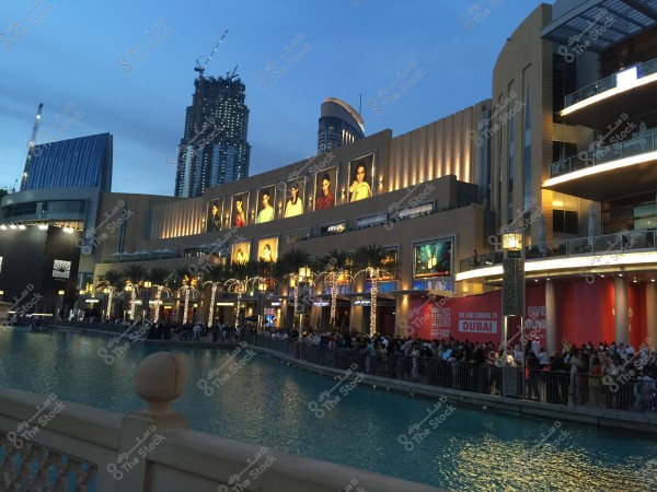 An image of a modern shopping mall facade in Dubai during the evening. The buildings are lit up with illuminated palm trees along the front. There is a crowd of people next to a calm blue lake. The architecture appears contemporary with large billboards displayed.