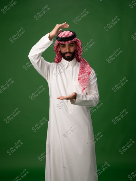 A portrait of a man standing against a green background wearing traditional Saudi attire, including a white thobe, a red and white ghutra, and a black agal. The man is gesturing with both hands, with his right hand above his head and his left hand in front of him, palm facing upwards.