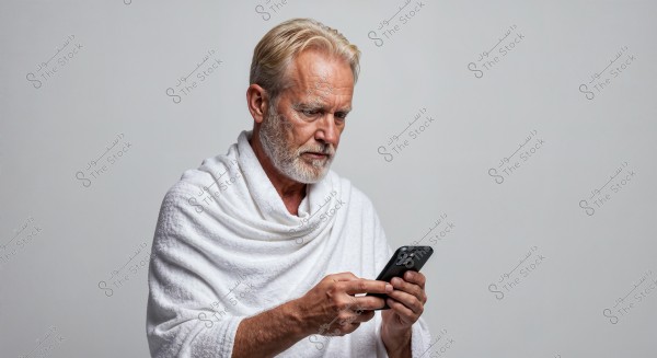 A portrait of an elderly man with white hair and beard, wearing a white robe resembling the attire used in Ihram. He is holding a mobile phone and appears focused on the screen. The background is plain with a light gray color.