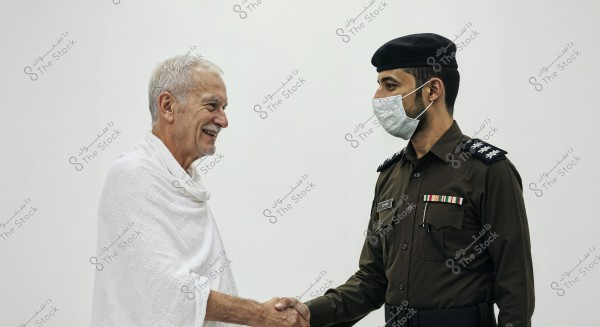 Two men shaking hands. The first man is wearing a white Ihram, which is a garment worn by Muslims during the Hajj or Umrah pilgrimage. The second man is a military officer wearing a brown uniform with rank insignia, a black cap, and a medical face mask. The background is plain white.