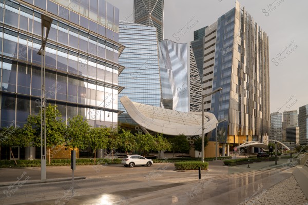 Image of the Financial District in Riyadh, Saudi Arabia. The picture showcases a collection of modern glass buildings with innovative architectural design. Some buildings reflect the sunlight, adding an attractive shine. In the foreground, trees and shrubs are neatly arranged along the road, with a white car driving down the street.