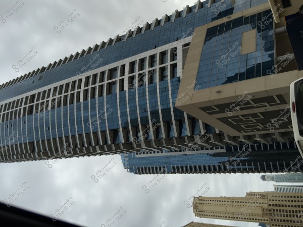 The image shows a modern skyscraper with contemporary architectural design. The building is covered with a glass facade that reflects the cloudy gray sky. The design features vertical and horizontal lines with a geometrically styled front facade and protruding sections. The skyscraper is surrounded by several other tall buildings, indicating an urban location.