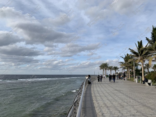 منظر جميل للواجهة البحرية يظهر المحيط على اليسار مع أمواج خفيفة وسور معدني يفصل البحر عن ممشى مرصوف واسع على اليمين. يتزين المسار بأشجار النخيل على الجانبين ومجموعة من الأشخاص يسيرون ويتمشون ويجلسون, بينما تبدو السماء مغطاة بسحب متوسطة وكثيفة. يبدو أن الموقع يستحضر إحساساً بالهدوء والراحة.