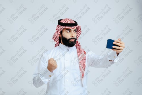 رجل سعودي يرتدي الزي الرسمي التقليدي على خلفية بيضاء, تصفح الهاتف المحمول, دردشه ومحادثات, التواصل مع الأصدقاء, مكالمات فيديو, القراءه من الهاتف المحمول, قضاء وقت ممتع وهادئ, تسليه وترفيه.