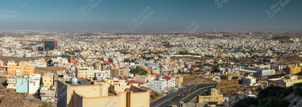 An image showcasing a panoramic view of an urban area in Saudi Arabia. The photo features numerous buildings in various colors and designs, with a major road intersecting through the city. The surrounding land and mountainous terrain are visible in the background under a clear blue sky.