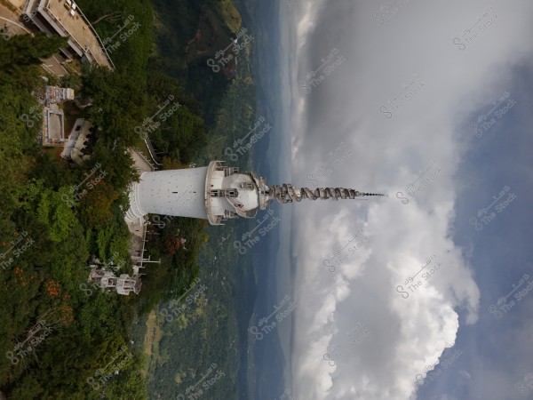 Image of a tall white radio tower in a lush mountainous area, surrounded by green trees. The tower features spiral elements with a building nearby. The sky is filled with clouds with some blue patches, and mountainous terrain is visible in the background.