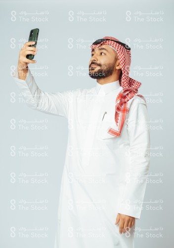 بورتريه رجل سعودي يرتدي الزي السعودي التقليدي يرفع الجوال لالتقاط السيلفي، شاب يأخذ صورة شخصية بالهاتف المحمول