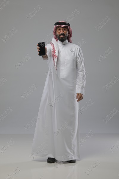 Image of a man wearing traditional Saudi attire, a white thobe, and a red and white shemagh on his head. He is smiling and extending his hand and foot forward, holding a smartphone in his right hand against a simple gray background.