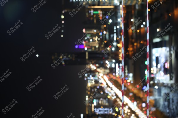 A nighttime image of a city featuring skyscraper lights and illuminated roads. The scene shows tall buildings scattered across the horizon with diverse lighting in colors such as blue, purple, and white. In the foreground, there are red and white barriers. The dark backdrop enhances the glow of the lights.