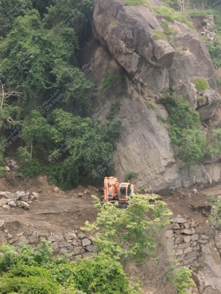 Image of a mountainous area covered with green plants and trees, dominated by a large rock formation. An orange excavator is seen performing earthwork near the base of the rock, while unevenly stacked stone walls are visible in the foreground. Green foliage is scattered throughout the scene, giving an impression of lush nature.