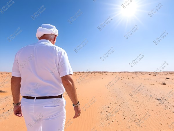 The image shows a person standing in the desert under the bright sun. The person is wearing traditional white clothing, including a shirt and trousers, with a head covering. The sky is blue and clear, and the sandy ground extends to the horizon.