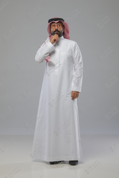 An image of a man wearing traditional Saudi attire, consisting of a white thobe, a red and white keffiyeh, and a black agal. The man is standing against a gray background, looking thoughtful with his hand on his chin.