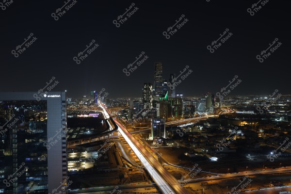 A night view of the Riyadh city skyline in Saudi Arabia. The image showcases the illuminated towers and skyscrapers, with the Kingdom Tower prominently visible with its distinctive lighting. Highways intersect in the foreground, displaying light trails from moving traffic across the city.