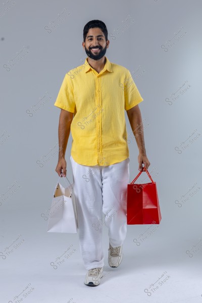 A man wearing a yellow shirt and white pants is holding two shopping bags, one white and one red, while smiling and looking at the camera. He is walking on a white floor with a simple gray background.