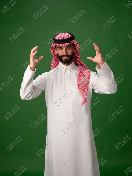 A photo of a man standing against a green background, wearing traditional Saudi attire. He is dressed in a white thobe and a red and white ghutra with a black agal. The man is raising his hands as if interacting with something unseen.