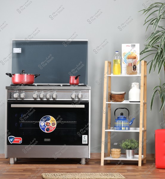 The image shows a modern kitchen with a silver stove featuring five knobs and four burners. On the stove, there are two red pots. Next to the stove is a wooden shelf with four levels containing a bottle of oil, a box of corn flakes, a blue teapot, and white and brown bowls, and a green potted plant. The floor is wooden with a small rug near the stove.