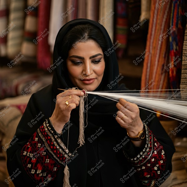 A woman wearing a black abaya and hijab with traditional embroidery, engaged in weaving using a needle and threads. The background displays colorful and embroidered fabrics. The woman is focused on her traditional work in what appears to be a handicraft shop.