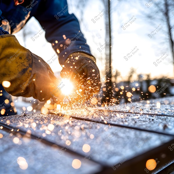 A close-up image of a person\'s hands wearing protective gloves during a welding operation outdoors. Sparks fly brightly in all directions, illuminated by a bright glow. The background features blurred outlines of trees and daylight, suggesting the work is being done in an outdoor setting during the day.