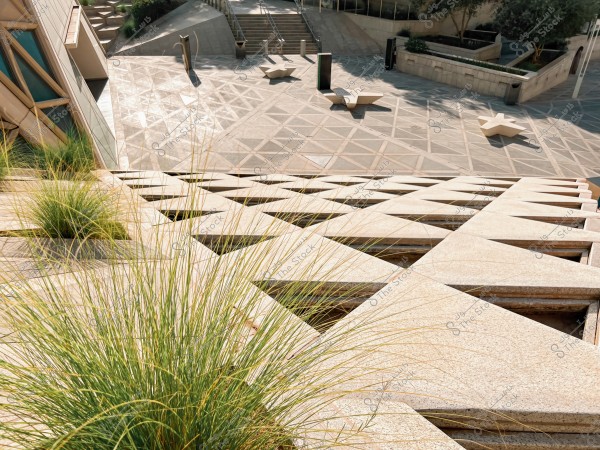 The image shows a modern geometric view of an outdoor area. The ground features repetitive triangular patterns, with grass plants extending along the left side surrounded by stone structures. In the background, star-shaped concrete seating and limited greenery are visible, with stairs leading to different levels. The area is sunny and clear within an urban setting.