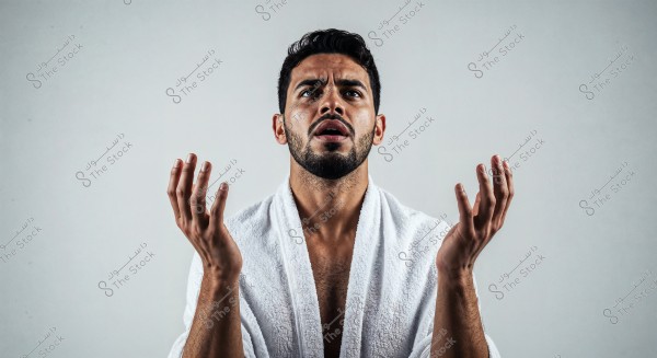 A close-up portrait of a man against a gray background, wearing a bright white robe resembling a bathrobe. His facial expression suggests focus or supplication, with his hands raised slightly forward.