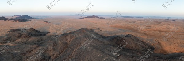 منظر لاحد المستطيلات الحجرية و صحراء واسعة مع جبال متفرقة. تظهر التضاريس القاحلة والمسطحات الرملية التي تمتد إلى الأفق تحت سماء صافية. الألوان الدافئة تميز الكثبان الرملية التي تندمج مع الجبال الصخرية الداكنة.