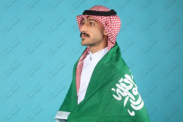 A young Saudi man wearing the Saudi uniform makes facial expressions and hands indicating the happiness and enthusiasm, Saudi flag