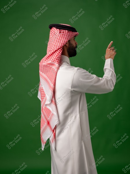 رجل يرتدي الثوب الخليجي التقليدي والشماغ يقف بجانب خلفية خضراء ويشير بإصبعه.