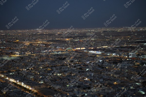 A night-time image of a large city illuminated by lights, showing numerous buildings, streets, and cars. In the center, there is a brightly lit bridge with vibrant colors. The sparkling lights extend across the horizon, highlighting the nightlife of the city.