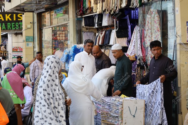 A photo of a group of people shopping in a busy outdoor market. Men are wearing traditional white thobes and kufis, while women are in hijabs and abayas. The merchandise includes colorful fabrics and clothes displayed on the wall. The scene appears crowded and local.