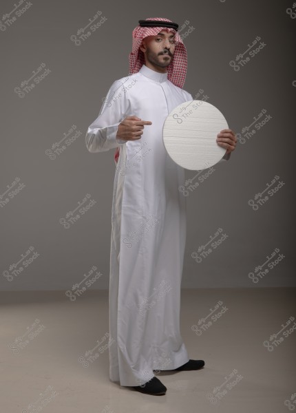 A man wearing traditional Saudi attire with a red and white shemagh and a black agal. He stands in the center of the image against a neutral grey background, holding a white circular disc in his left hand, and pointing at it with his right finger.