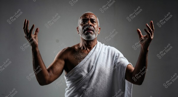 A photo of a man standing in front of a gray background, wearing a white cloth wrapped around his body similar to Hajj Ihram attire. He appears to be gesturing or raising his hands in an expressive manner. The man has a short white beard and short gray hair.