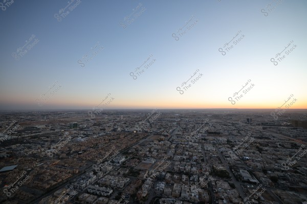منظر جوي لمدينة كبيرة تحت سماء صافية وقت الغروب, حيث يمكن رؤية المباني السكنية والتجارية جنباً إلى جنب مع شبكة من الطرق والشوارع. الأفق يظهر بوضوح مع تلاشي ضوء الشمس في الأفق, مما يضفي ألوان الشفق على السماء.