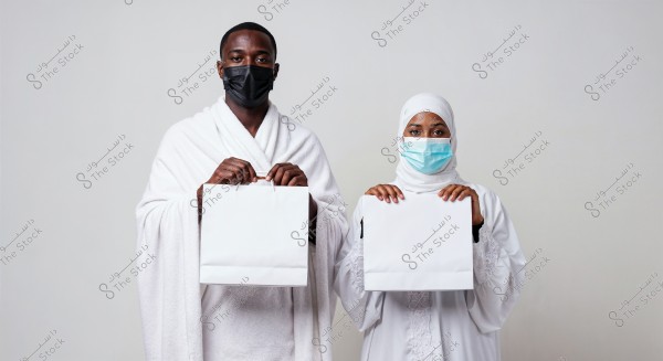 The image shows a man and a woman standing side by side, both wearing traditional white Ihram clothing and medical face masks. Each is holding a white shopping bag. The man appears to be wearing an izar and a shawl, while the woman is wearing an abaya and hijab. The background is plain and white.